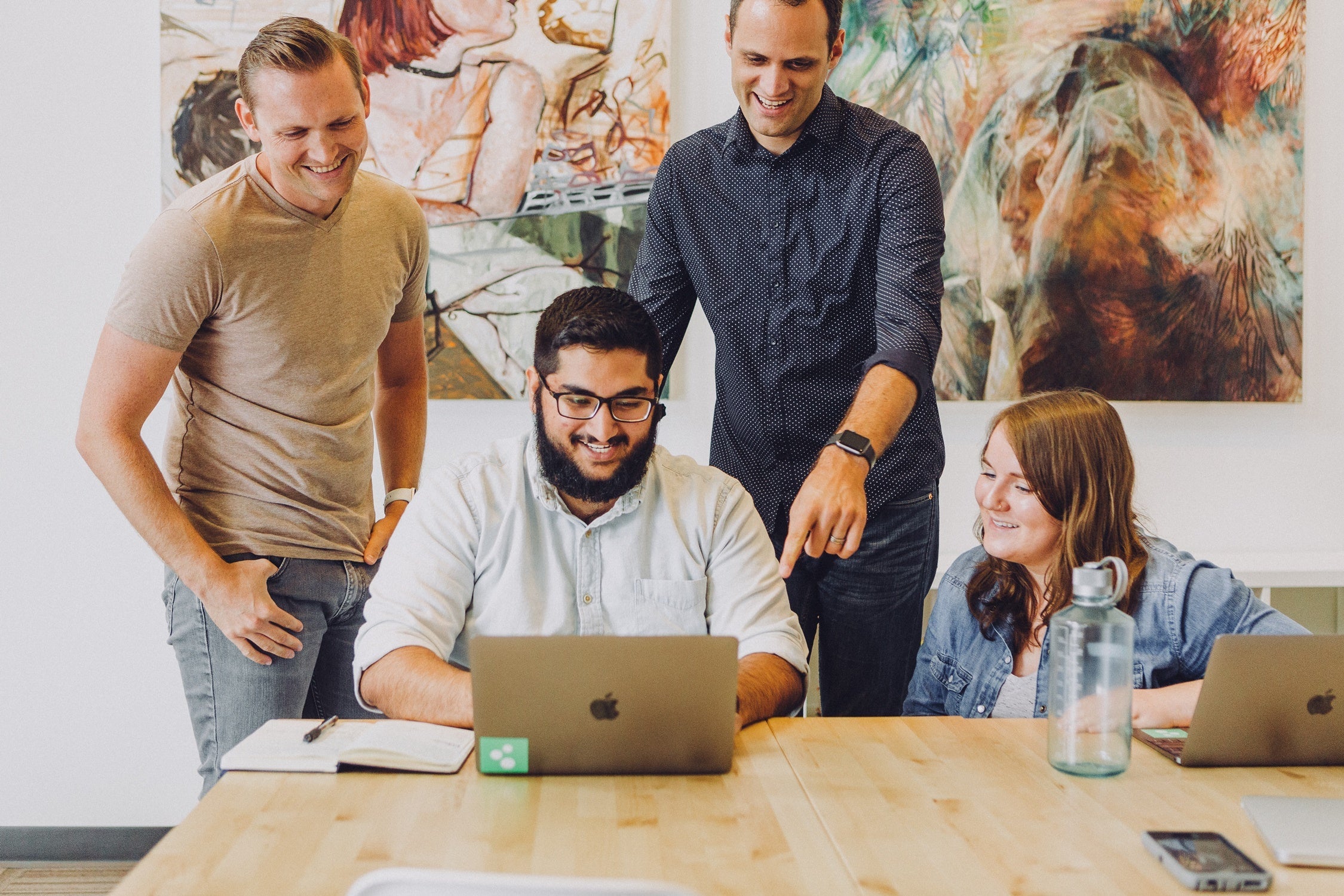 A group of people in an office gathering around a laptop with an abstract painting in the background.