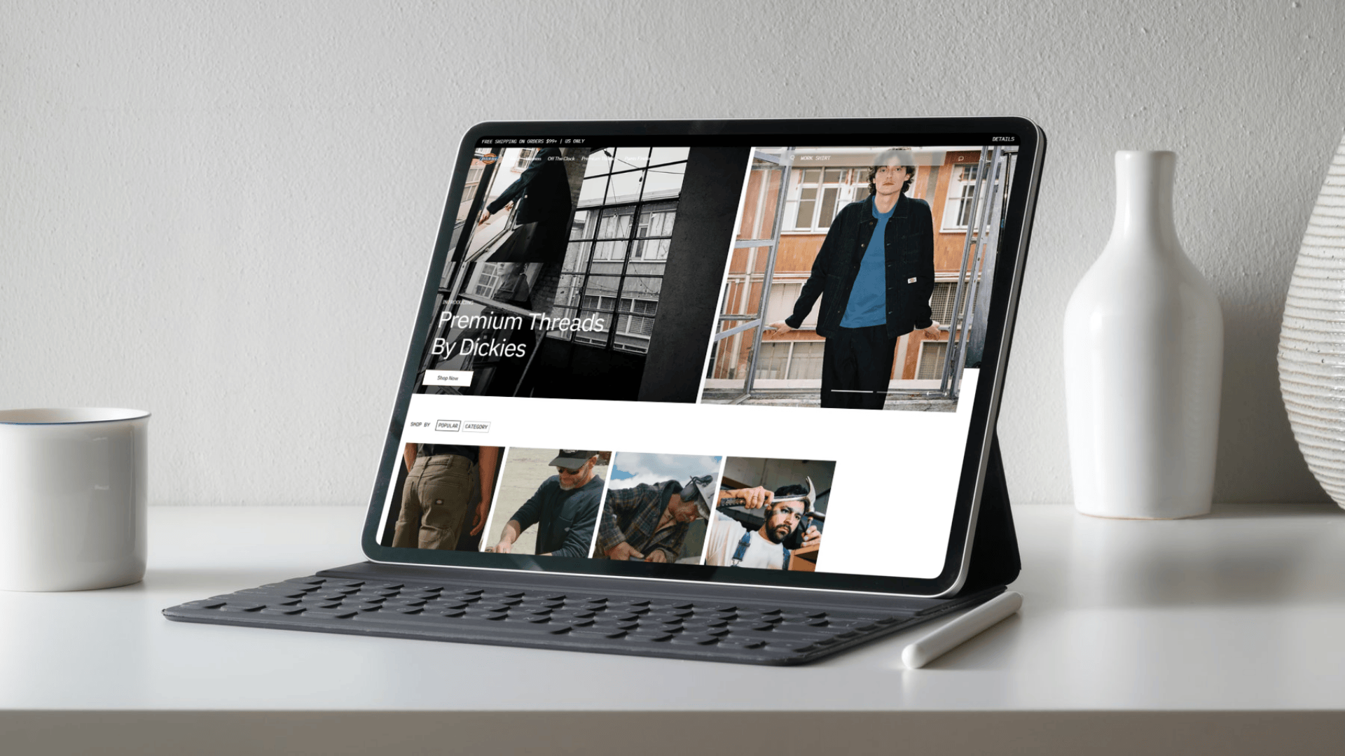 Tablet displaying a fashion website on a white surface with a vase and mug in the background