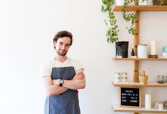 A smiling man wearing an apron standing with his arms crossed over his chest by a shelf.