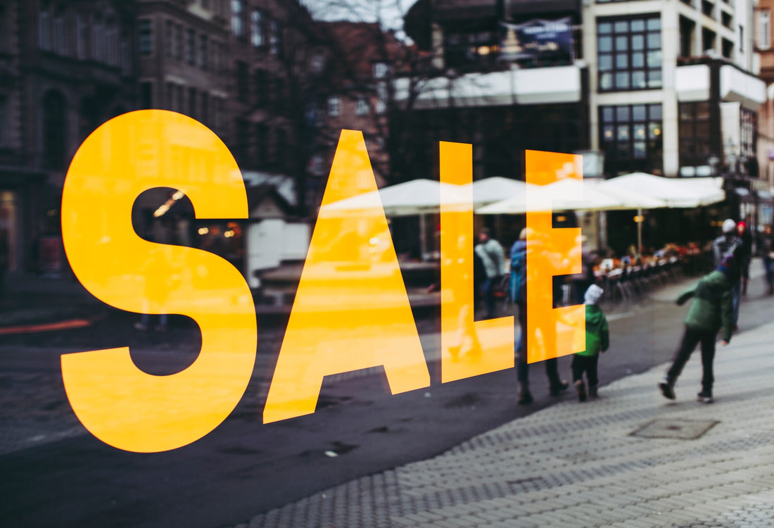 A SALE sign in yellow displayed on a glass wall reflecting an urban sidewalk.