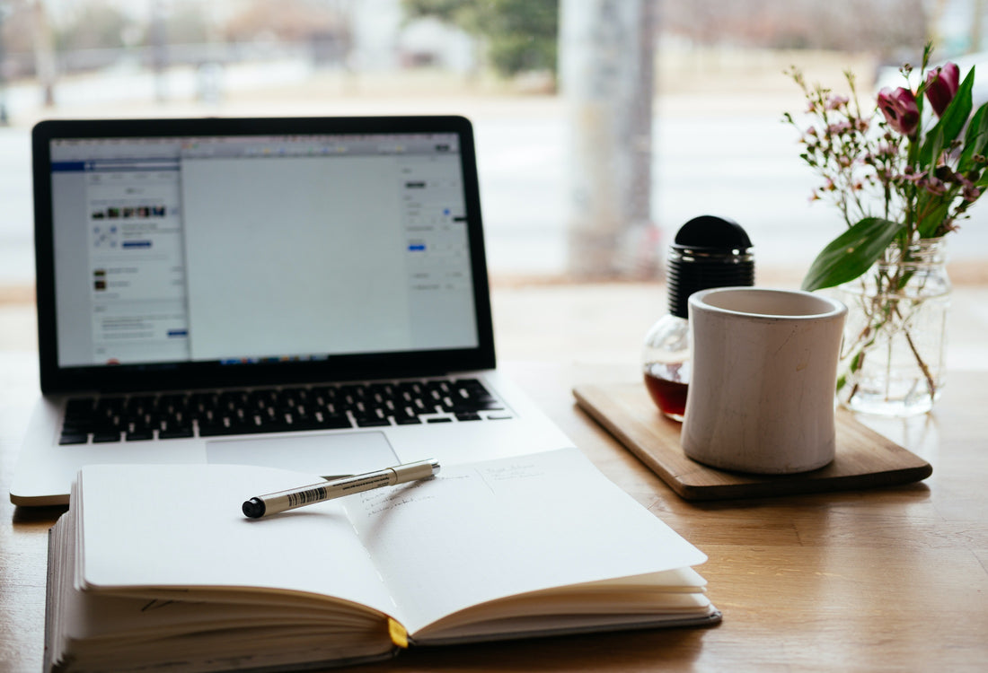 A laptop, open notebook, pencil, mug, and flower vase on top of a wooden table.