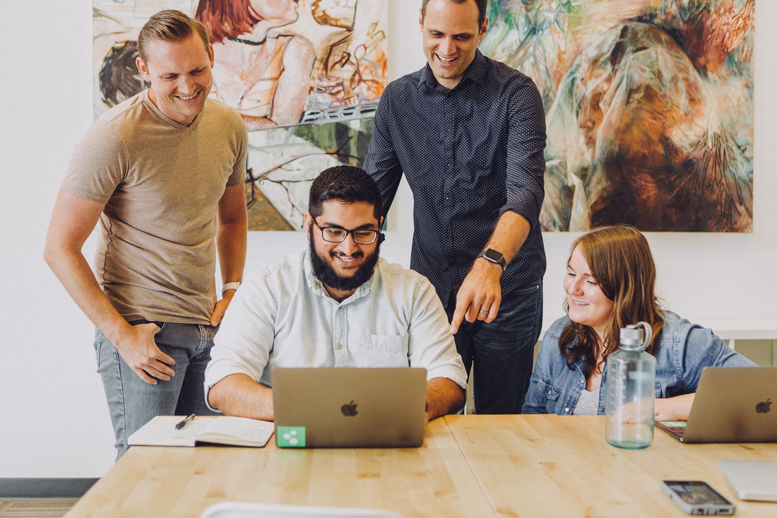 A group of people in an office gathering around a laptop with an abstract painting in the background. 