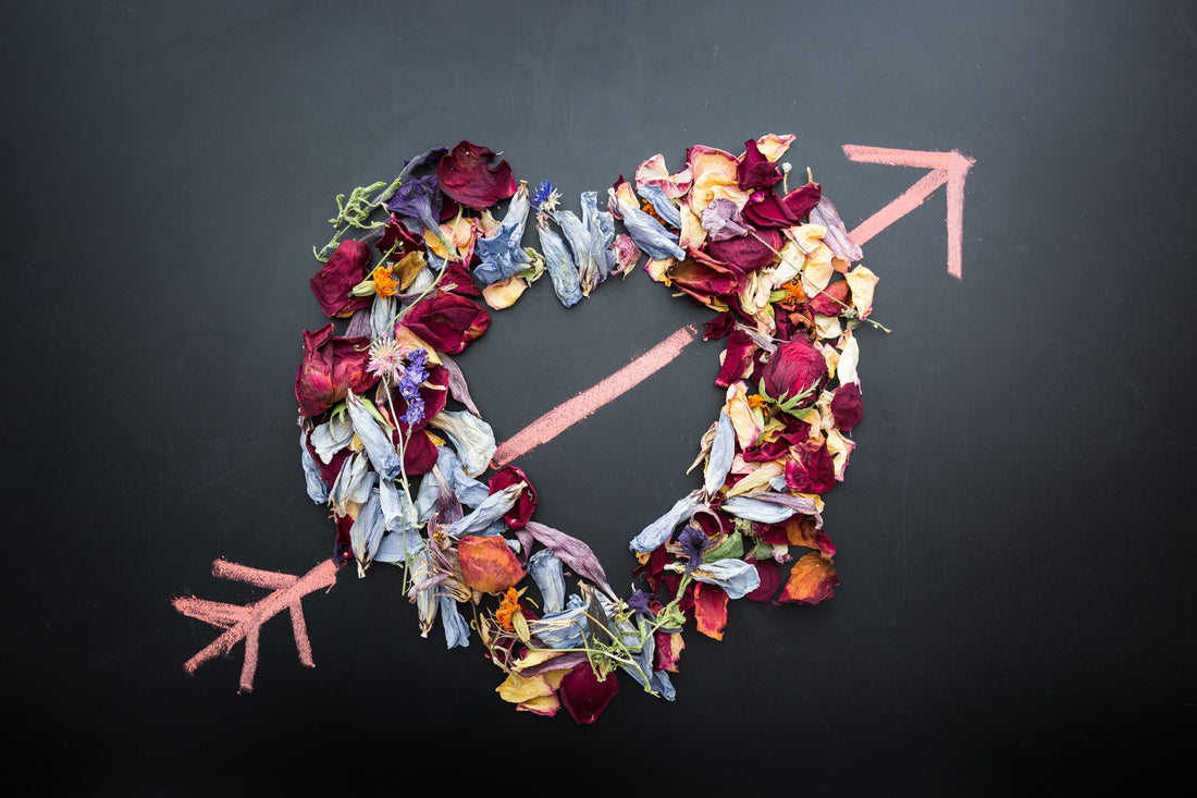 A flat lay of various colorful flower petals arranged in the shape of a heart with cupid's arrow going through it.