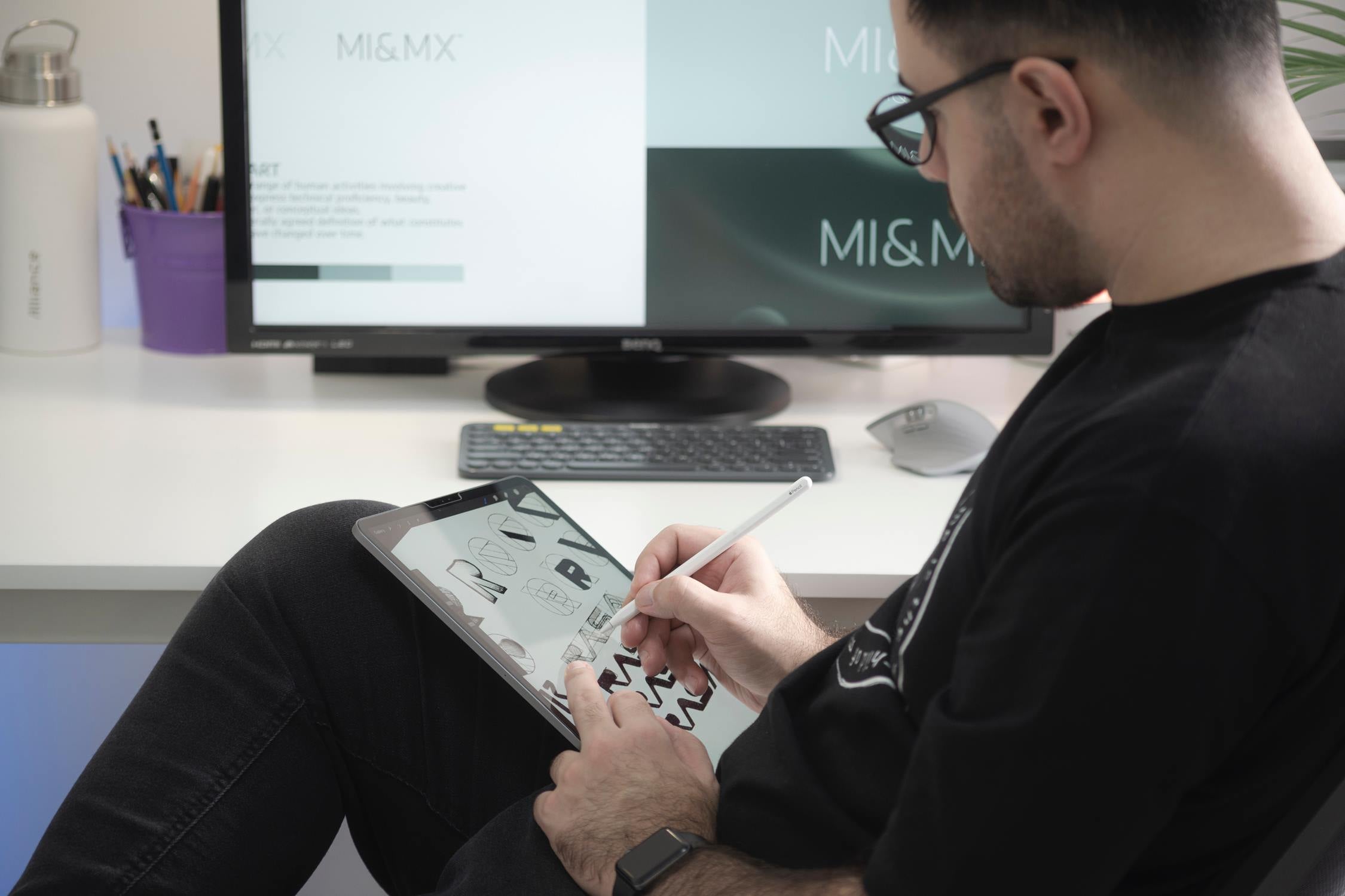 A man sketching on an iPad with a desktop computer in the background.