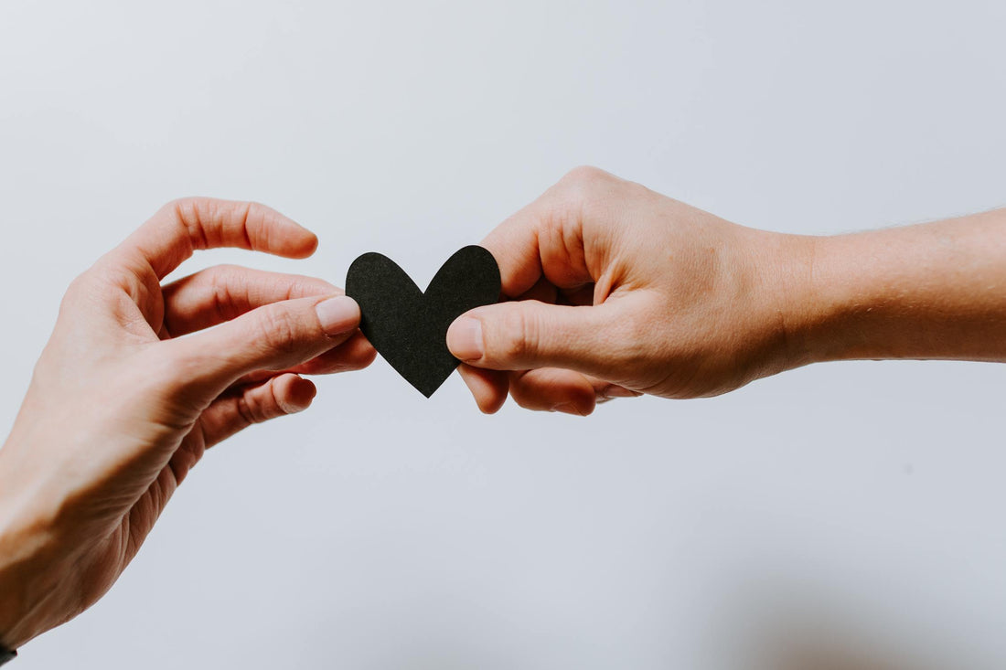 Two hands passing a black paper heart with a white background.