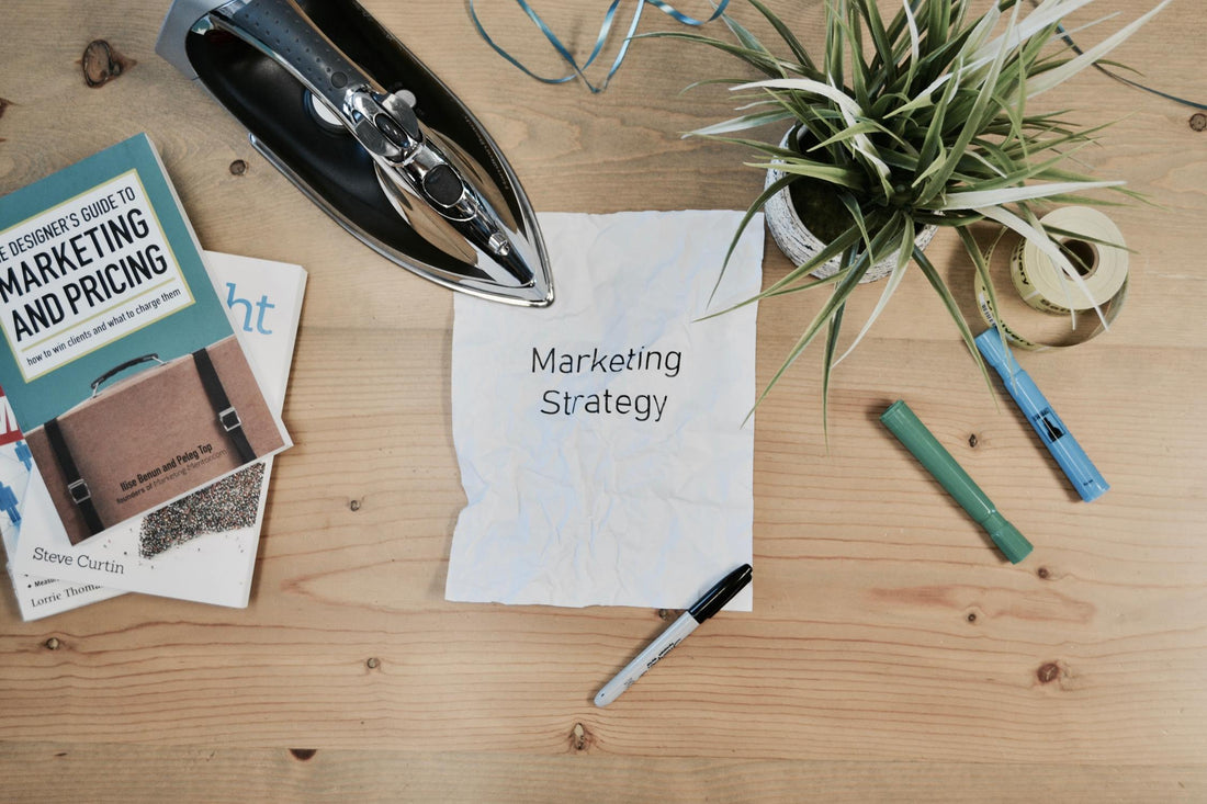 A flat lay of books, pens, a potted plant, iron, and crumped paper with Marketing Strategy printed across it.
