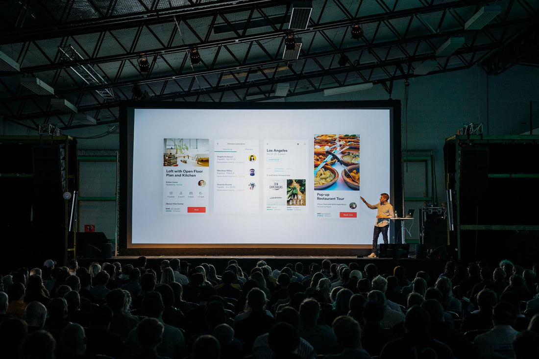 A theater-style venue full of people with a large projected image and speaker.