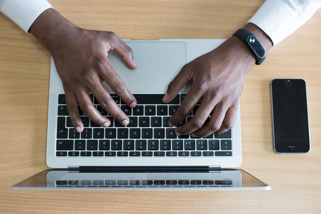A flat lay of two hands typing on a laptop with a phone on the right side of the image.