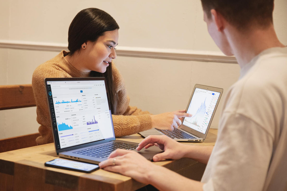 Two people with two laptops sitting on a desk discussing graphs on their laptop screens.