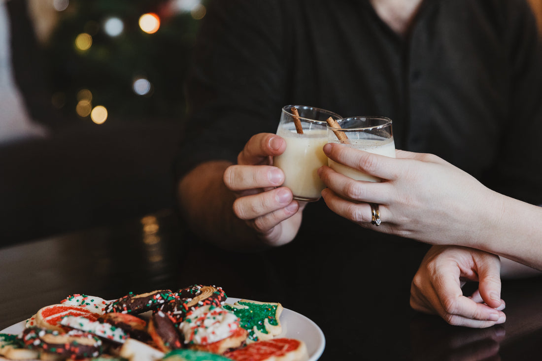 Two hands clinking glasses of eggnog over a plate of Christmas-themed cookies.
