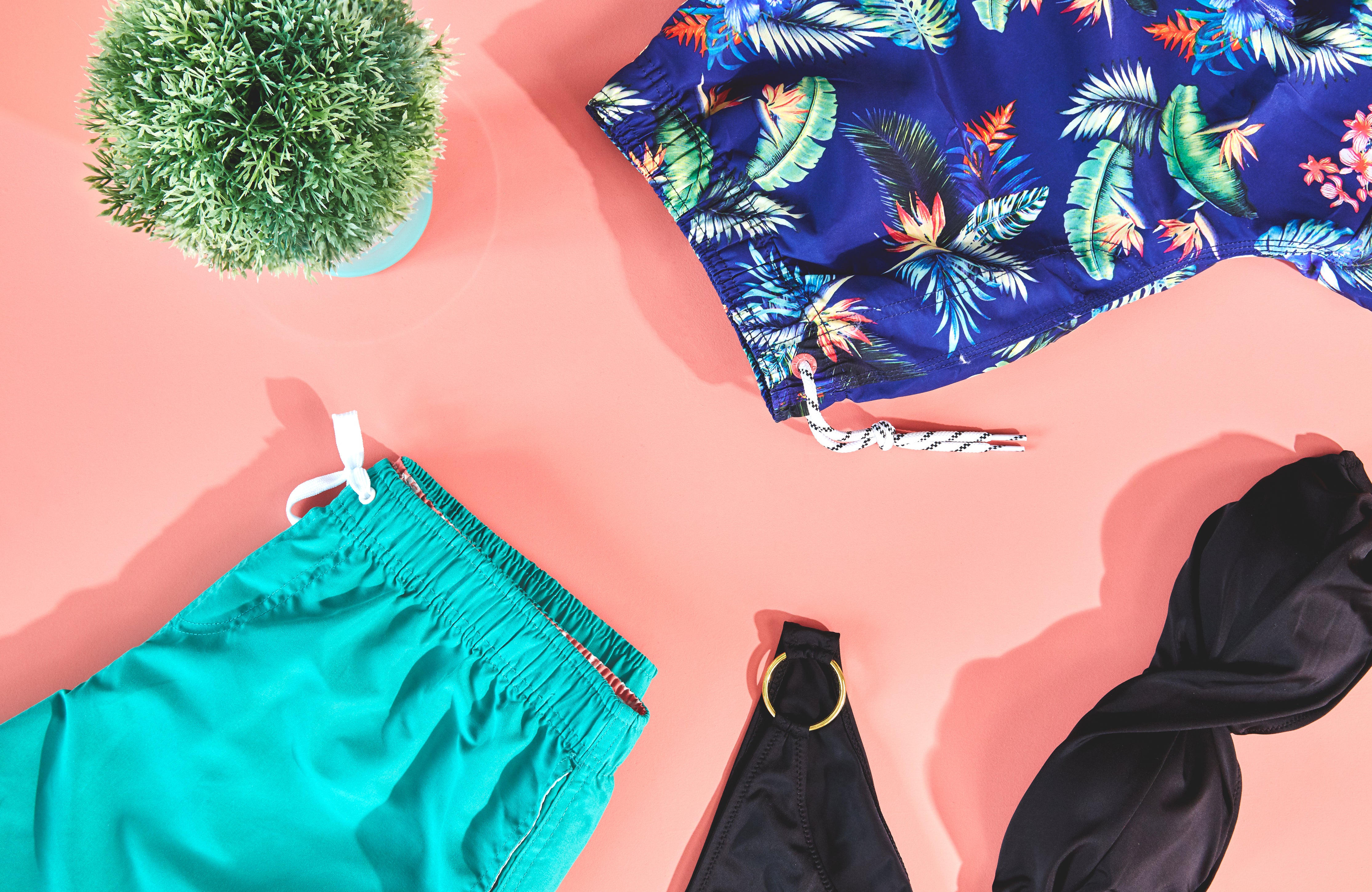 A summer-themed flat lay of a black bikini and two pairs of swimming shorts.