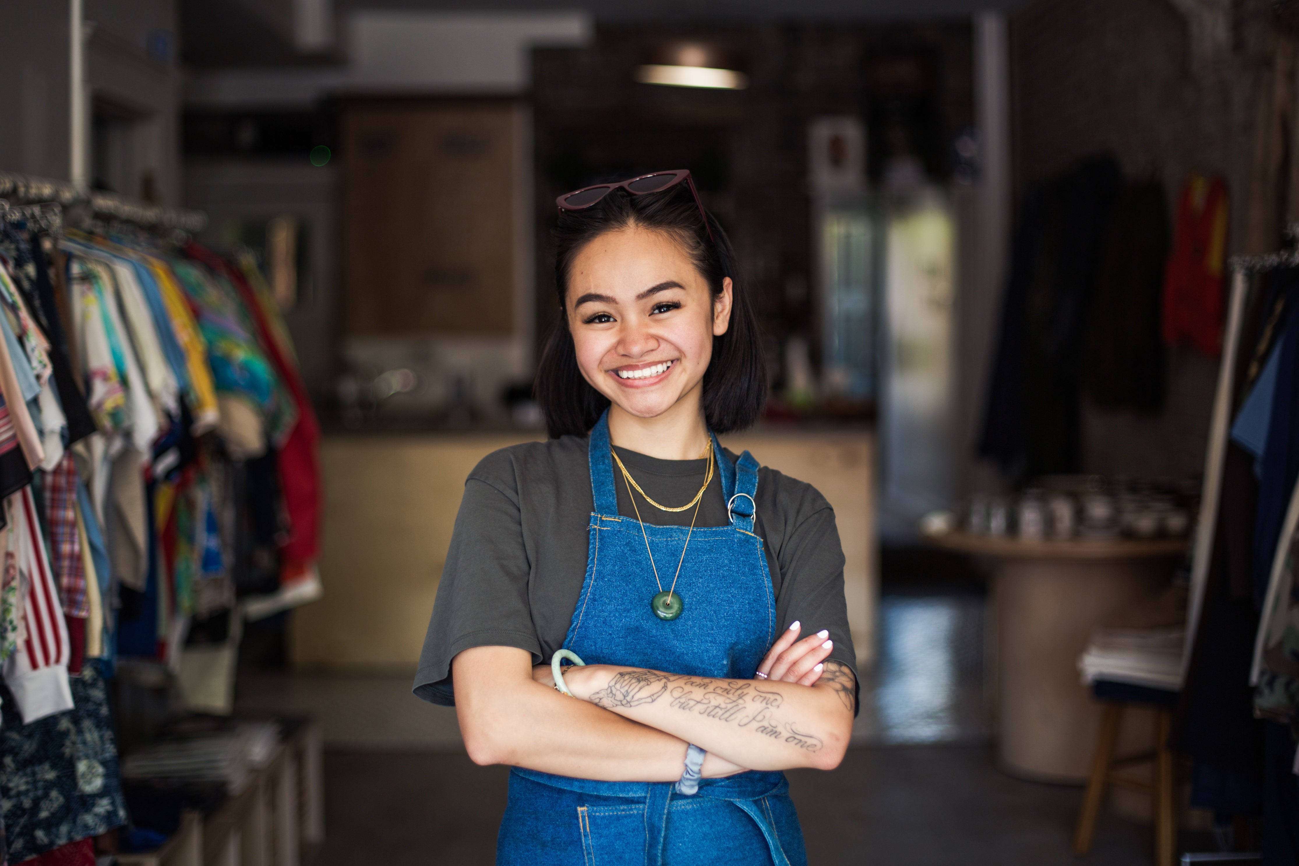 A smiling woman wearing denim overalls with her arms crossed over her chest standing inside a clothing store.